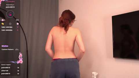 Media: Video of a topless woman with long brown hair in a ponytail, wearing only gray sweatpants, standing in a living room with a TV on the wall and a window covered by a gray curtain.