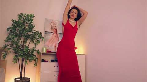 Media: Video of a woman with medium skin tone and dark hair, wearing a red dress, stretching her arms above her head in a room with a white wall, green plant, and a framed painting.