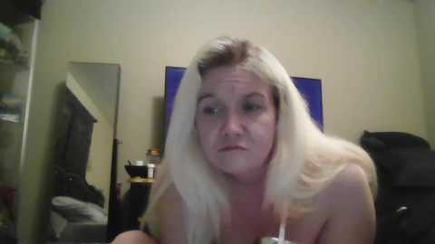 Media: Video of a Caucasian woman with long, platinum blonde hair, wearing a white tank top, sitting in a cluttered bedroom with beige walls and a mirror.