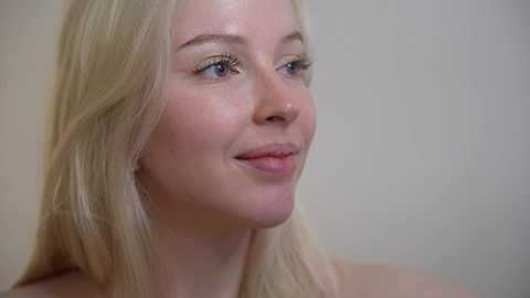 Media: A video of a fair-skinned woman with long, straight blonde hair, blue eyes, and light makeup, smiling softly, with a blurred, light-colored background.