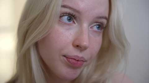 Media: Video of a fair-skinned, blonde-haired woman with blue eyes, slightly freckled cheeks, and a neutral expression. She wears light makeup and is indoors with a soft-focus background.