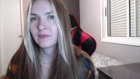 Media: Video of a young Caucasian woman with long, straight, blonde-streaked hair, wearing a plaid shirt, sitting at a desk in a modern room with a red and black gaming chair and white blinds.