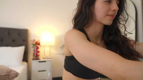 Media: Video of a young woman with long dark hair, fair skin, and wearing a black bra, standing in a modern bedroom with a white bed, gray headboard, and a lamp with red flowers.