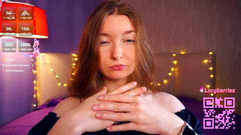 Media: Video of a light-skinned woman with long brown hair, wearing a black top, sitting on a bed with a headboard, surrounded by yellow string lights.