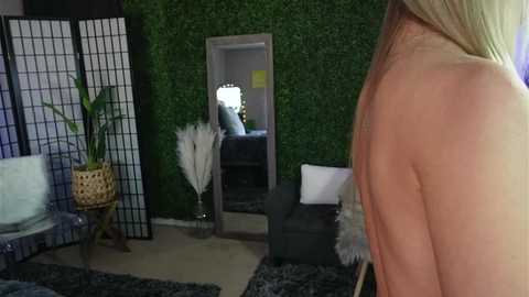 Media: Video of a woman's bare back, blonde hair, in a modern living room with a green wall, black couch, and a potted plant.