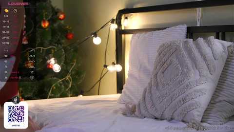 Media: Video of a cozy bedroom with a bed featuring white pillows and a knitted blanket. A Christmas tree adorned with lights and ornaments is visible in the background, creating a warm, festive atmosphere.