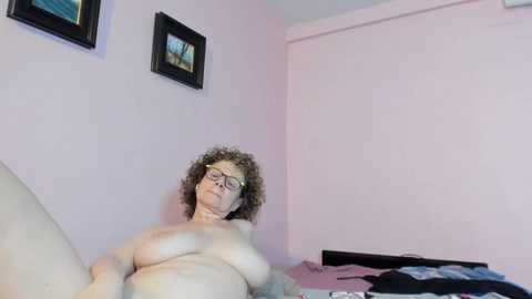 Media: A video of a nude, older woman with curly hair and glasses, lying on a bed in a pale pink room with framed art on the wall.