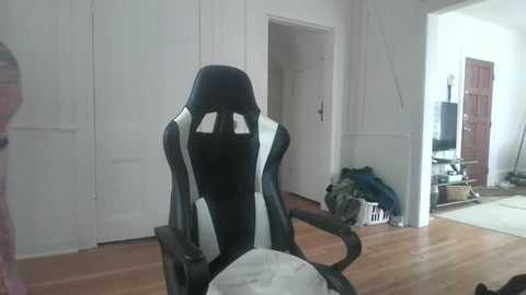 Media: A video of a sleek black and white gaming chair with a high backrest in a minimalist, white-walled room with hardwood floors, a door open to a cluttered hallway.