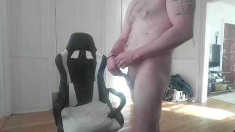 Media: Video of a nude, slender, fair-skinned man holding his erect penis, standing in front of a black and white gaming chair in a dimly lit room with hardwood floors.