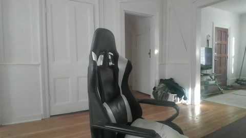 Media: Video of a modern gaming chair with a black and white design, positioned in a bright, minimalist room with white paneled walls, wooden floor, and open doorways leading to other rooms.