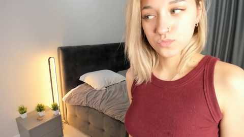 Media: Video of a blonde woman with a nose ring, wearing a red ribbed tank top, standing in a modern bedroom with a grey upholstered bed and potted plants on a nightstand.