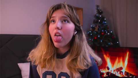 Media: Video of a blonde teenage girl with long hair, sticking out her tongue, wearing a navy and beige sports jersey, in a cozy living room with a lit Christmas tree and a roaring fireplace.