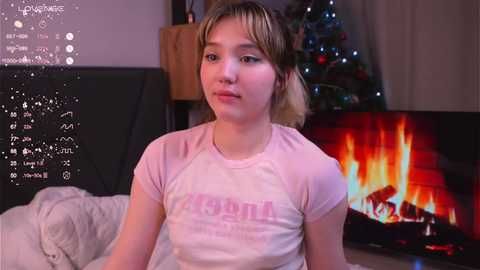 Media: Video of a young woman with light skin and short blonde hair, wearing a pink t-shirt with text, sitting in front of a lit fireplace, surrounded by a cozy room with a Christmas tree in the background.