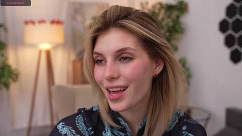 Media: Video of a smiling, light-skinned woman with long, straight blonde hair, wearing a black and blue patterned blouse, standing indoors next to a lit table lamp and a potted plant.