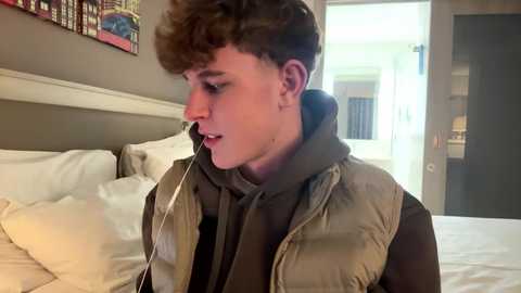 Media: Video of a young man with curly brown hair, wearing a puffy vest and headphones, sitting on a bed in a modern bedroom with beige walls and a colorful abstract painting.