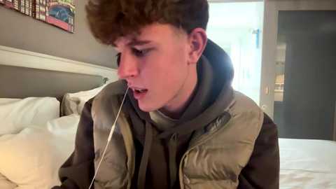 Media: Video of a teenage boy with curly brown hair, wearing a puffy green vest over a black hoodie, listening to earphones in a modern bedroom with white sheets and a colorful wall art.