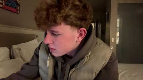 Media: Video of a young man with curly brown hair, wearing a gray puffer vest, leaning on a white bed, looking contemplative, in a dimly lit, beige-walled bedroom.