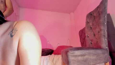 Media: Video of a partially nude woman with fair skin and a small tattoo on her shoulder, leaning against a tufted, dark grey upholstered headboard in a pink-lit bedroom.