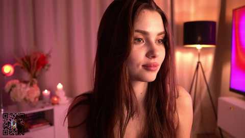 Media: Video of a young woman with long brown hair, light skin, and a slender figure, smiling softly. She is in a warmly lit, softly decorated room with a tall lamp and flowers in the background.