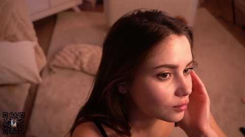 Media: A video of a young woman with long brown hair, fair skin, and a contemplative expression, touching her cheek. She is in a cozy room with beige carpeting, a bed, and soft lighting.