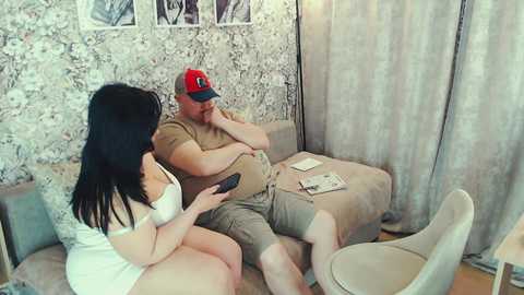 Media: Video of a young woman with long black hair in a white dress, sitting on a beige couch with a man in a red cap and brown shirt, both looking at a smartphone.