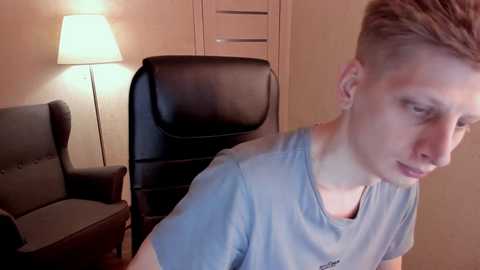 Media: Video of a young man with short, light brown hair, wearing a light blue T-shirt, sitting in a dark leather office chair in a dimly-lit room with a beige wall and a lamp beside him.