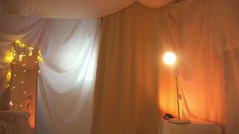 Video of a warmly lit, simple room with white curtains, soft orange light, a round table with a lamp, and fairy lights in the background.