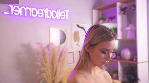 Video of a nude, fair-skinned, blonde woman with medium build, standing in a softly lit room with minimalist decor, including a \"Tempted\" neon sign, a potted plant, and a shelf with white vases.