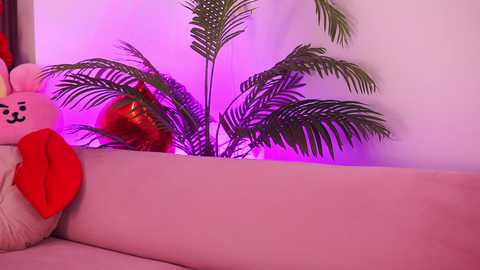 Media: Video of a plush, pink, cartoonish teddy bear with a red bow, lying on a white couch, with a potted palm plant and purple lighting in the background.