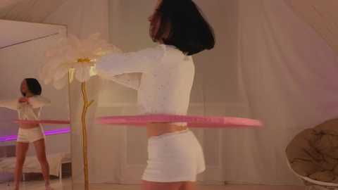 Media: Video of a woman in a white crop top and mini skirt, twirling a pink hoop, in a softly lit, tent-like room with mirrors and white decor.