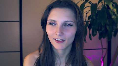 Media: Video of a young Caucasian woman with long brown hair, fair skin, and blue eyes smiling, wearing a strapless top, standing against a backdrop of a green plant and a beige, folding screen.