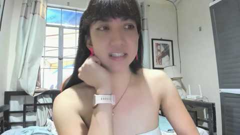 Media: Video of an Asian woman with long black hair, wearing a white strapless top and a smartwatch, lying on a bed in a dimly lit room with a window and framed picture on the wall.