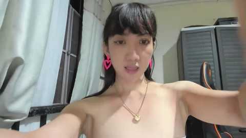 Media: A young Asian woman with straight black hair, wearing pink heart-shaped earrings, and a gold necklace, taking a selfie, topless, in a dimly lit room with beige walls and a window.