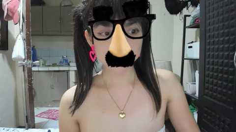 Media: Video of a young Asian woman in a bathroom with black hair, wearing oversized black glasses, pink earplugs, and a black beak mask. She has a necklace and a pink towel nearby.