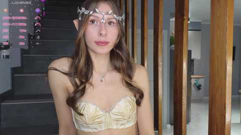 Media: Video of a young woman with fair skin, long brown hair, and elf ears, wearing a gold lace bra and a jeweled headband. She stands in a modern, dimly lit room with wooden doors and a staircase in the background.