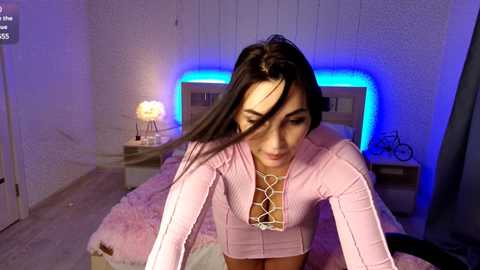 Media: Video of a young woman with long dark hair, wearing a pink ribbed top with a lace-up front, sitting on a bed with a pink blanket. The room has white brick walls and a blue neon light strip behind her.