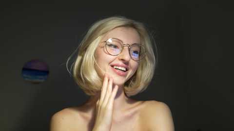 Media: A video of a smiling, light-skinned blonde woman with shoulder-length hair and round glasses, wearing nothing, with her hands near her face. The background is dark with a hint of a blue and purple abstract painting.