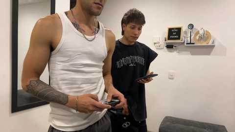 Media: Video of a muscular man with tattoos in a white tank top and a young boy in a black t-shirt, both using smartphones in a minimalist room with white walls and a shelf.