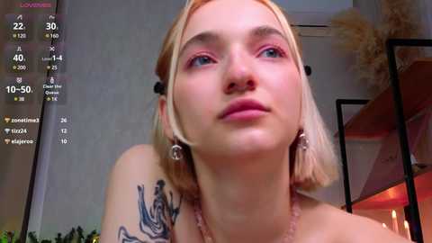 Media: Video of a young, fair-skinned woman with platinum blonde hair and pink eyeshadow, wearing a black top and dangling earrings. She has a tattoo on her shoulder. Background shows a modern room with a TV screen displaying a live stream.