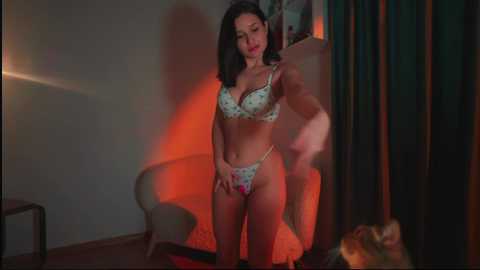 Media: Video of a slim Asian woman with shoulder-length black hair in floral lingerie, adjusting her bra strap. Background features a dimly lit room with a glass table, green curtains, and a chair.