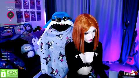 Media: A video of a young woman with straight, bright orange hair in a black crop top, seated next to a plush shark costume. The room features posters, plush toys, and a blue curtain background.