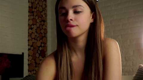 Media: Video of a young, light-skinned woman with long brown hair, wearing a white top, standing indoors. Background features a patterned wall and a bed with a wooden headboard.