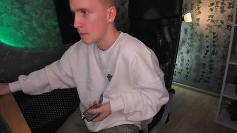 Media: A video of a young man with short blonde hair, wearing a white sweatshirt and gray pants, seated at a desk in a dimly lit room with green and black walls.