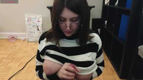 Video of a fair-skinned woman with shoulder-length dark hair, wearing glasses, and a black-and-white striped sweater, exposing her large breasts, seated on a chair in a room with wooden floors, a pink rolling chair, and bookshelves.