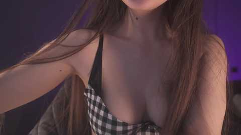 Media: Video of a light-skinned woman with long brown hair, wearing a black-and-white checkered top that reveals cleavage, against a purple backdrop.