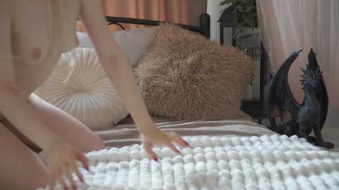 Media: Video of a fair-skinned, nude woman with small breasts, arranging white, round balls on a bed with beige pillows and a fluffy cushion, alongside a black metal headboard and a small dragon figurine.