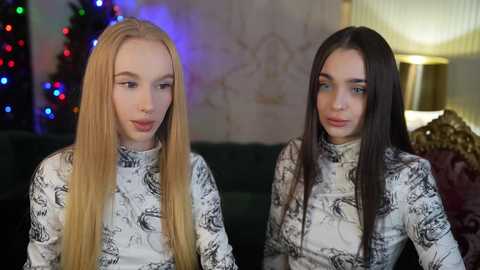 Media: Video of two young women with long hair, one blonde and one brunette, wearing white patterned tops, sitting side-by-side on a dark couch in a cozy, festive room with a lit Christmas tree and warm lighting.