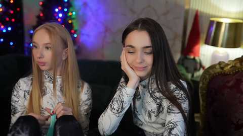 Media: Video of two young women, one blonde with long hair, the other brunette, both with closed eyes and hands on faces, sitting on a dark couch. Background includes a lit Christmas tree, a red flag, and a lamp.