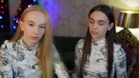 Media: Video of two young women with long hair, one blonde and one brunette, in white patterned sweaters, sitting indoors with a blurred festive background.