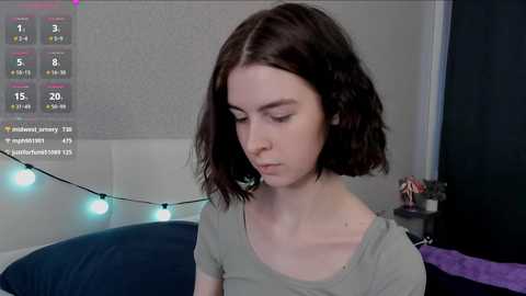 A video of a young woman with shoulder-length dark hair, wearing a light green top, sitting indoors. Background includes a white headboard, a blue pillow, and a string of fairy lights.